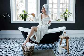 A cute young woman wearing a white robe and having a towel on her head spend time in the bathroom