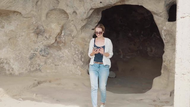 cute young woman with blond hair in summer light clothes and dark sunglasses, tourist comes out of cave in Georgian mountains and calls friend with phone, smiles cheerfully and shares her impressions