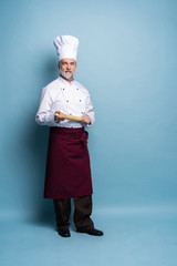 Full length of mature professional chef with rolling pin on blue background.