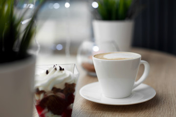 White cup of hot cappuccino on white saucer and red velvet dessert on wooden bar table next to window. Cold snowy weather outside