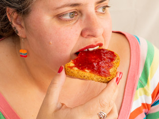 An adult woman with red nails and lips eating a rusk with raspberry jam