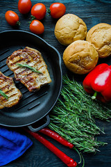Grilled steak on a round grill pan, garnished with spices for meat, rosemary, greens and vegetables on a dark wooden background.