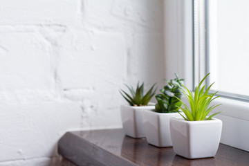 The green plastic cactus is in a white pots. On a brown window sill against a white brick wall.