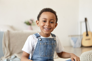 Fototapeta premium Indoor shot of handsome cheerful happy black little boy of school age posing in blurred interior with guitar in background, smiling joyfully at camera, playing alone while his parents at work
