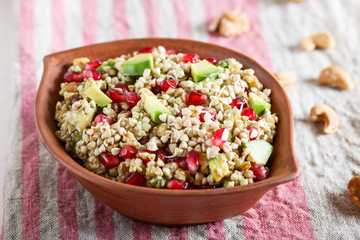 Salad of germinated buckwheat, avocado, walnut and pomegranate seeds in clay plate on white wooden background.