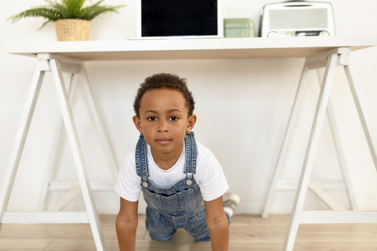 Picture of handsome cute Afro American boy of preschool age posing in stylish apartment interior, looking at camera with curious or suspicious look, being home alone. Selective focus on face