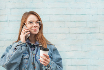 Attractive girl student talks on the phone on the background of a blue wall, holds a paper cup with coffee looking in the direction and smiles, wearing a jeans jacket. Copyspace