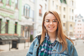 Portrait of a cheerful girl wearing casual clothing, standing in headphones, background of the landscape of the old town,looking camera and smiling.Close portrait of a call girl is listening to music