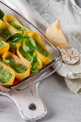 Cooking vegetarian pasta recipe with greens, herbs, tomatoes and cheese, baked in glass tray on white wooden background.