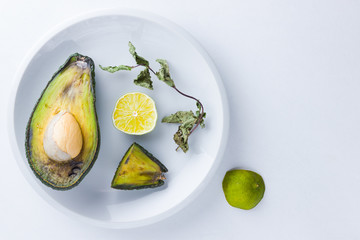 Ugly food on white background. Ugly rotten avocado on white plate. Bad lime and dry mint on a plate. Rotten tropical fruits. Concept of rotten fruit. Top view, copy space