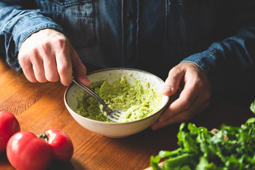 Man mashing avocado in bowl, wooden table background. Food preparation image, healthy vegan and vegetarian food