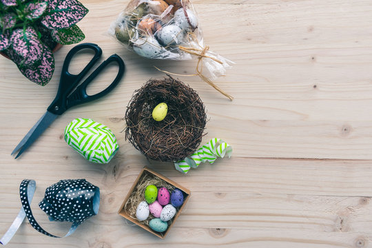Top view of accessories or equipment of making a colorful Easter eggs nest with scissors, a green plant, colorful quail eggs decorations, ribbons and a straw nest on pale wooden background.