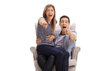 Happy young couple in an armchair pointing at the camera and laughing