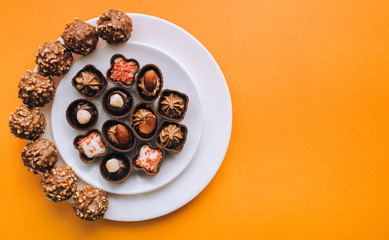 Beautiful chocolates of different shapes and fillings lie in a white plate against the background of orange paper. Top view, copy space.