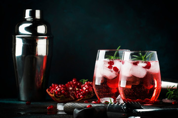 Red cocktail with cold vodka and pomegranate juice, ice cubes and rosemary, bar tools, blue black...
