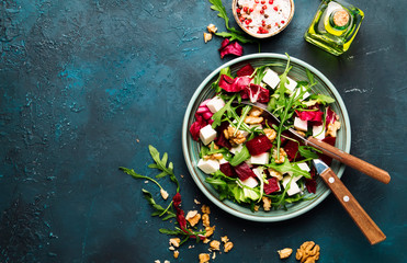 Beet or beetroot salad with fresh arugula, radicchio, soft cheese and walnuts on plate with fork, dressing and spices on blue kitchen table background, copy space, top view