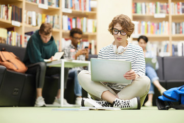Serious pensive curly-haired student girl with headphones on neck sitting with crossed legs on floor in library and analyzing online resources on laptop