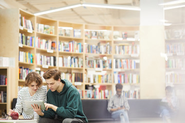 Serious concentrated young students in casual clothing sitting in modern library area and using tablet while searching for useful information together