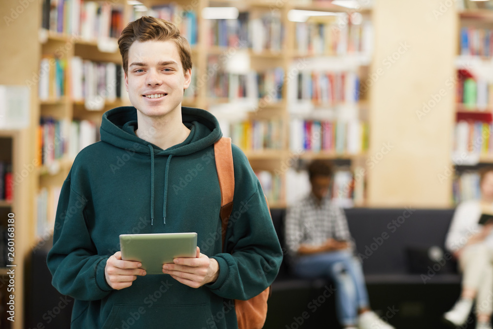 Wall mural Cheerful confident handsome high school student with satchel standing in modern library and using digital tablet while looking at camera