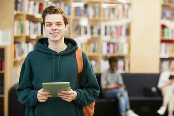Cheerful confident handsome high school student with satchel standing in modern library and using digital tablet while looking at camera