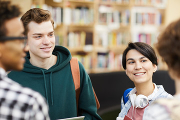 Group of positive confident young students in casual clothing standing in circle and chatting with each other in university library