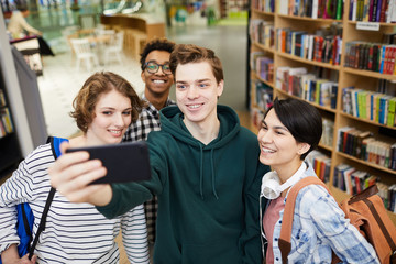 Group of cheerful excited young multiethnic students in casual clothing standing in book store and taking selfie together on smartphone