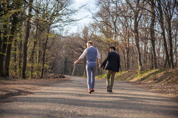 rear view, two gay man walking in forest, on asphalt road.