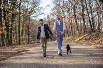 Fototapeta premium front view, two gay man walking in forest, on asphalt road. Together holding hands.