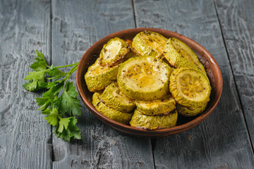Pieces of baked zucchini in a clay bowl with herbs on a wooden table.