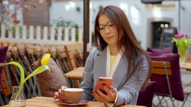 charming asian woman is drinking coffee in open terrace in cafe in spring day, using smartphone