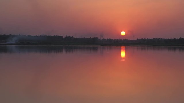 Sun setting in a brilliant colorful red and orange sky with reflection in the water across the lake and forest somewhere on the highway in Karnataka, India 