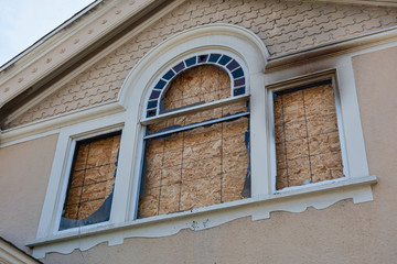 Home window boarded up with plywood sheets. House fire - walls and eves charred.  Plywood shutters prevent unauthorized access by squatters, looters to damaged unused, vacant, or abandoned property.