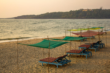 empty blue beach beds under color awnings on the yellow sand against the background of the sea and the green island with the house in the evening sunset