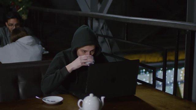 Tracking left of young hooded man sitting in caf&eacute; at laptop, typing and drinking tea