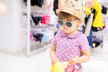 Adorable smiling little girl at the clothes store, try on straw hat and sunglasses for summer