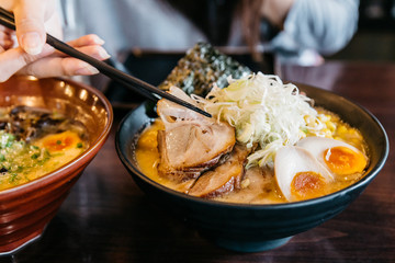 Woman hand pinching noodle in Ramen Pork Bone Soup (Tonkotsu Ramen) with Chashu Pork, Scallion, Sprout, Corn, Dried Seaweed and boiled egg served in black bowl.