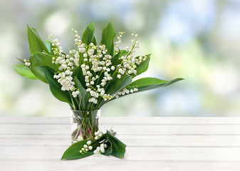 Bouquet of flowers and leaves lily of the valley ( Convallaria majalis ) in small glass on white wooden table on blur garden background