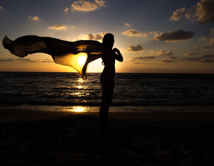 A girl stands on the beach at sunset and holds a shawl that flies in the wind.