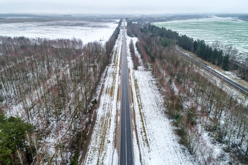 Fototapeta premium The road passing through the fields, on the sides of the road is growing forest. Fields covered with snow.