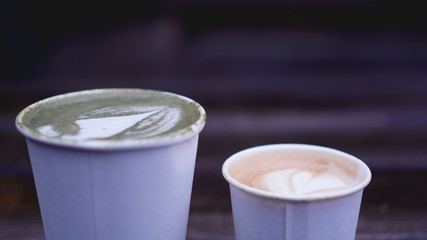 Cup of matcha tea with latte art and cappuccino in paper cup on wooden background. Blurred background.