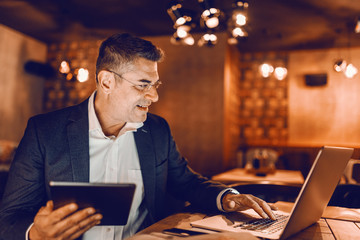 Profile of middle aged businessman dressed smart casual typing on laptop and holding tablet while sitting in cafeteria.