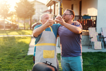 Father in law and his son in law making a toast with beer and hugging while standing next to grill. Family gathering concept.