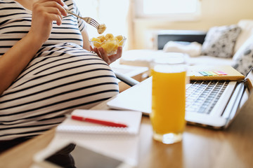Young Caucasian pregnant freelancer eating fruits and sitting at home office. On desk laptop, glass with fresh orange juice and notebooks. Working pregnant women concept.