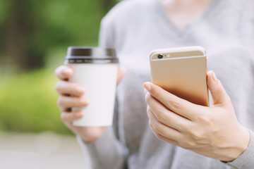 Young woman lady attractive chatting on her mobile smartphone, tablet reading text message on cell while relaxing in the public park, hold hand coffee cup standing drinking coffee with in cozy morning