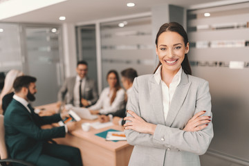 Beautiful Caucasian brunette in formal wear standing with arms crossed and smiling. In background colleagues having meeting. You don't build a business. 