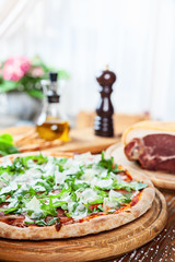 Fresh pizza with prosciutto on a wooden table on light background. Olive oil, pepper, prosciutto and flowers on background. Vertical image with copy space for design. Italian cuisine. Food Lunch time