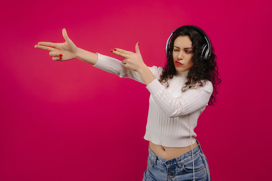 Beautiful girl showing shooting geisture listening to music with headphones isolated on pink background