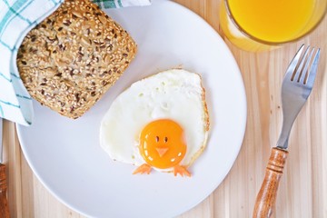 A fried egg lies on a plate, next to it are bread rolls and cutlery. The egg yolk of the fried ice cream looks like a chick, with two grains of eyes and pieces of carrot as mouth and feet