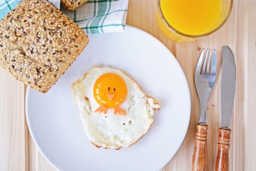 A fried egg lies on a plate, next to it are bread rolls and cutlery. The egg yolk of the fried ice cream looks like a chick, with two grains of eyes and pieces of carrot as mouth and feet