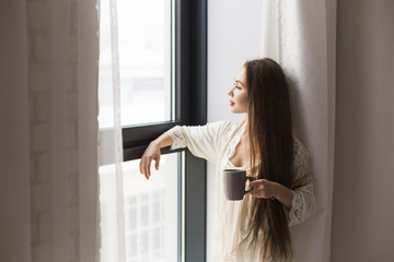 Attractive young girl in night clothes standing by the window with a mug of coffee